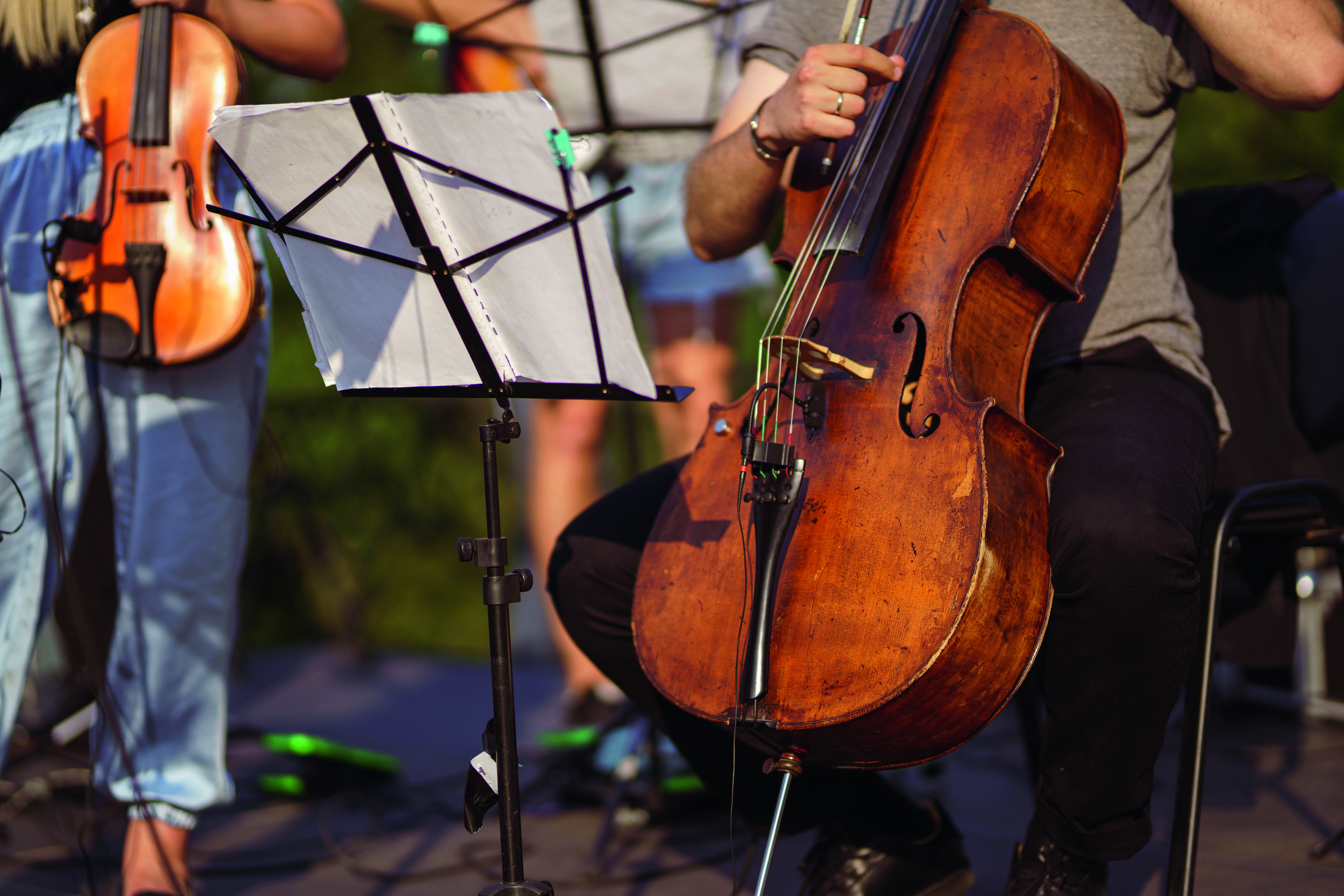 Close up of man cellist sitting on chair near music stand with notes and holding violoncello while having concert rehearsal on the street